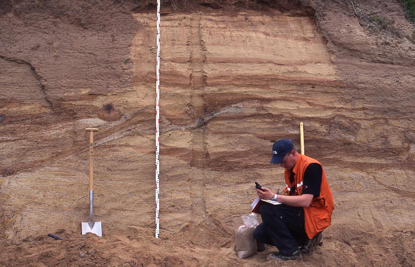 Rotbraune Steinbruchwand, davor ein Geologe in Warnweste und mehreren Arbeitsutensilien