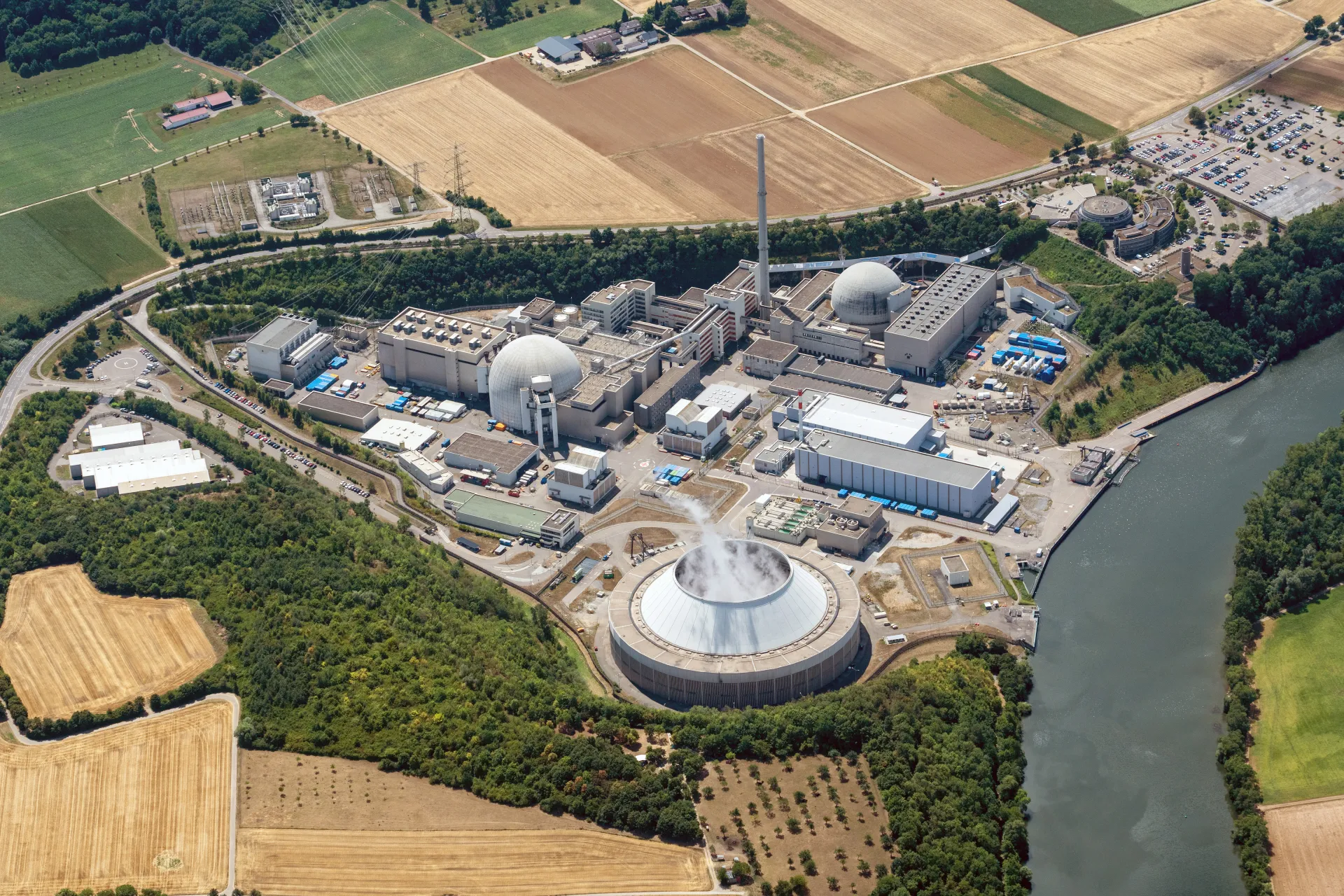 Blick auf einen Gebäudekomplex in strahlendem Sonnenschein aus der Vogelperspektive. Etwa mittig in der unteren Bildhälfte ist ein dampfender Kühlturm zu erkennen. Am rechten Bildrand verläuft ein Fluss unmittelbar entlang der Anlage. Der Gebäudekomplex wird von Hecken und schmalen Wäldern umschlossen. In der weiteren Umgebung sind Felder und vereinzelte Bauernhöfe zu erkennen.