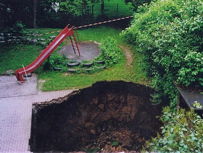 Ansicht eines Erdfalls im Hof eines Kindergartens. Unmittelbar angrenzend zu dem steilen, runden Loch in der Wiese bzw. des gepflasterten Hofs befindet sich ein Kinderspielplatz mit Rutsche.