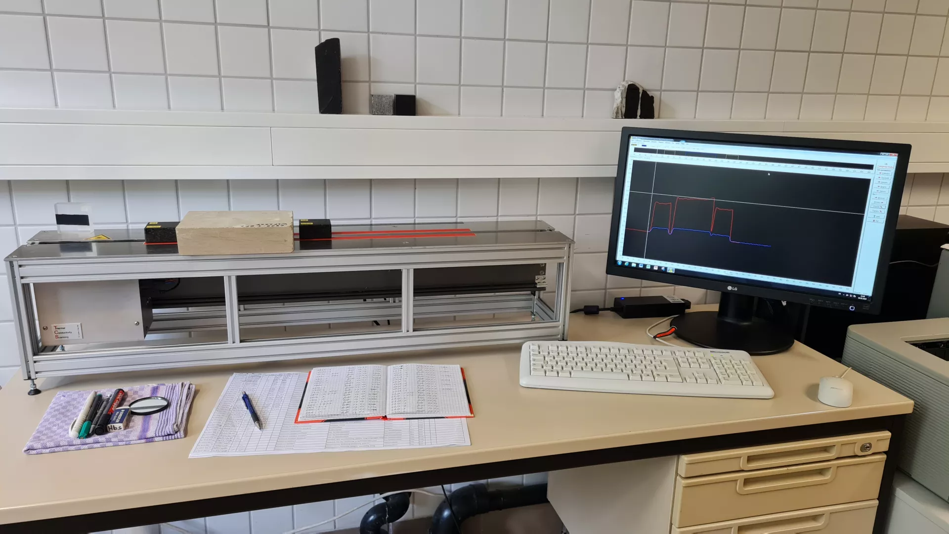Das Foto zeigt ein etwa ein Meter langes und 30 Zentimeter hohes Laborgerät, welches auf einem Schreibtisch steht. Mit dem Gerät kann die Wärmeleitfähigkeit von Festgesteinen gemessen werden. An das Gerät ist ein Computer mit Bildschirm und Tastatur angeschlossen.