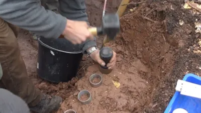 In einer in rötlichem Tonboden aufgegrabenen Profilgrube werden rundliche Metallzylinder eingeschlagen, zur Entnahme von Bodenproben.