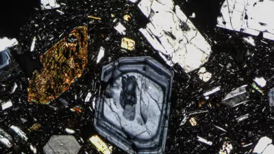 Das Foto zeigt einen Gesteinsdünnschliff, aufgenommen unter einem Polarisationsmikroskop. Das Gestein ist ein Vulkanit mit großen Mineralen in feinkörniger Matrix. An manchen Mineralen ist ein Zonarbau (Kristallwachstum mit unterschiedlichem Chemismus) erkennbar.