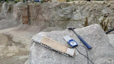 Auf einem grauen Granitblock liegen  ein Meterstab, ein Kompass und ein Hammer. Im Hintergrund ist ein Steinbruch zu sehen.