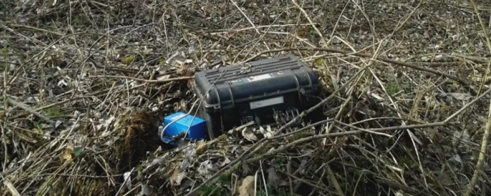 Auf dem Boden liegende Äste und Laub. In der Mitte eine temporäre Erdbebenmessstation mit einem blauen Seismometer und einer schwarzen Box für die Geräte der Messdatenspeicherung