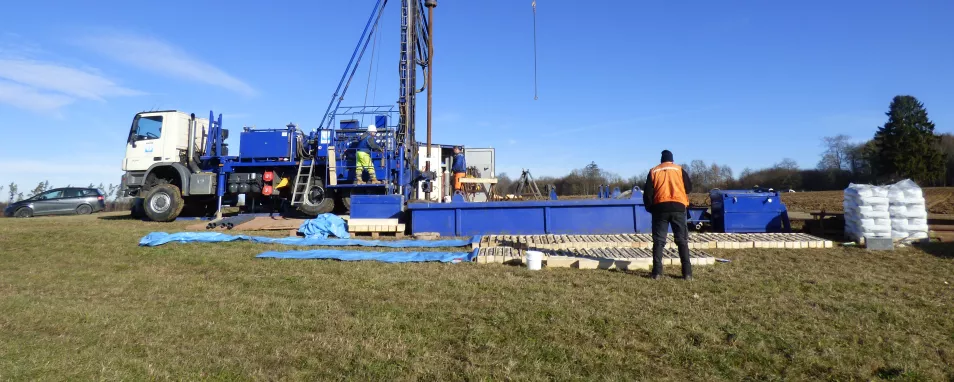 Blick auf einen blauweißen LKW mit montiertem Bohrturm vor blauem Himmel auf einer Wiese. Zwei Personen bedienen das Bohrgerät. In der unteren Bildhälfte sind schmale Holzkisten mit Bohrkernen ausgelegt. Eine Person in oranger Weste in Rückenansicht steht vor den Holzkisten und betrachtet diese.