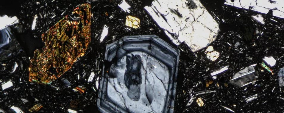 Das Foto zeigt einen Gesteinsdünnschliff, aufgenommen unter einem Polarisationsmikroskop. Das Gestein ist ein Vulkanit mit großen Mineralen in feinkörniger Matrix. An manchen Mineralen ist ein Zonarbau (Kristallwachstum mit unterschiedlichem Chemismus) erkennbar.